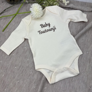 May include: A cream-coloured baby onesie with long sleeves. The text "Baby Toutounji" is printed in brown script across the chest. The onesie has snap closures at the bottom and a round neckline. The garment is made of soft material.
