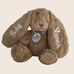 Puede incluir: Un conejo de peluche marrón con orejas largas y caídas. La oreja izquierda está bordada con la palabra Baby en letra blanca, y la oreja derecha presenta el nombre Stanes en letra blanca. Una etiqueta redonda con un logo de koala cuelga del cuello.
