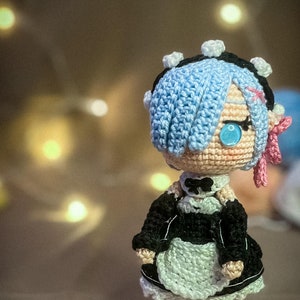 May include: A handmade crochet doll of a character with blue hair and blue eyes. The doll is wearing a black and white maid outfit with a frilly apron. The doll has a pink bow and is set against a blurred background.