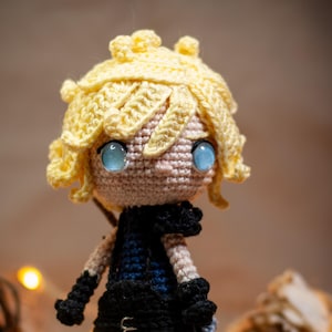Puede incluir: Una muñeca de crochet con cabello amarillo, ojos azules y un atuendo negro y azul. La muñeca está de pie sobre una superficie marrón con luces de cadena en el fondo.