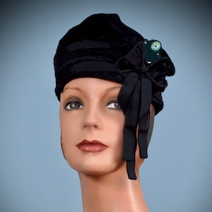 May include: A black velvet hat with a large black bow and a green jewel accent. The hat is worn on a mannequin's head.