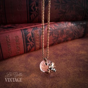 May include: A gold-toned box chain necklace featuring a clear, spherical glass pendant adorned with a small, metallic bee charm. The bee has textured wings and a dark, patterned body, suspended directly from the chain alongside the glass sphere.