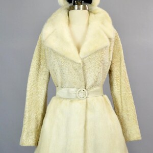 May include: A white fur coat with a matching fur hat and a white satin belt. The coat has a collar and a button closure.