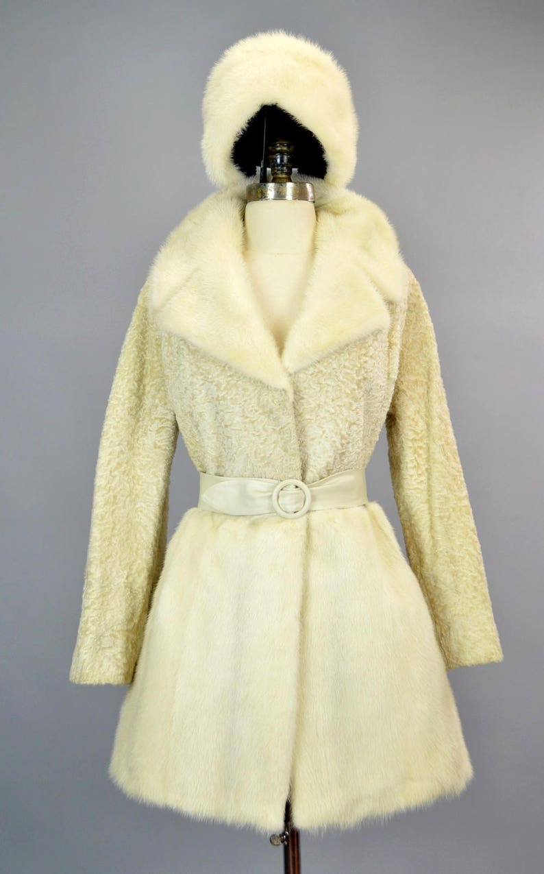 May include: A white fur coat with a matching fur hat and a white satin belt. The coat has a collar and a button closure.