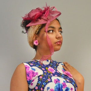 Peut inclure: Une femme portant une robe à fleurs rose et blanche avec un chapeau rose et des boucles d'oreilles assorties. Le chapeau a une plume rose et un ruban rose.