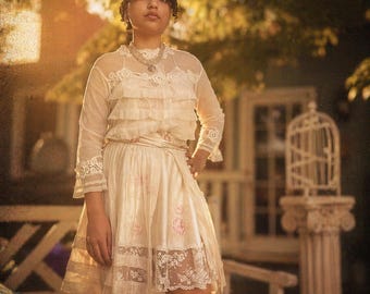 May include: A cream-coloured vintage dress with lace and ruffle details. The dress features long sleeves with lace cuffs, a tiered bodice, and a skirt with floral accents and a lace hem. A necklace is worn around the neck.