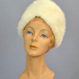 May include: A white fur hat with a wide brim. The hat is worn on a mannequin with a light brown face and blue eyes.
