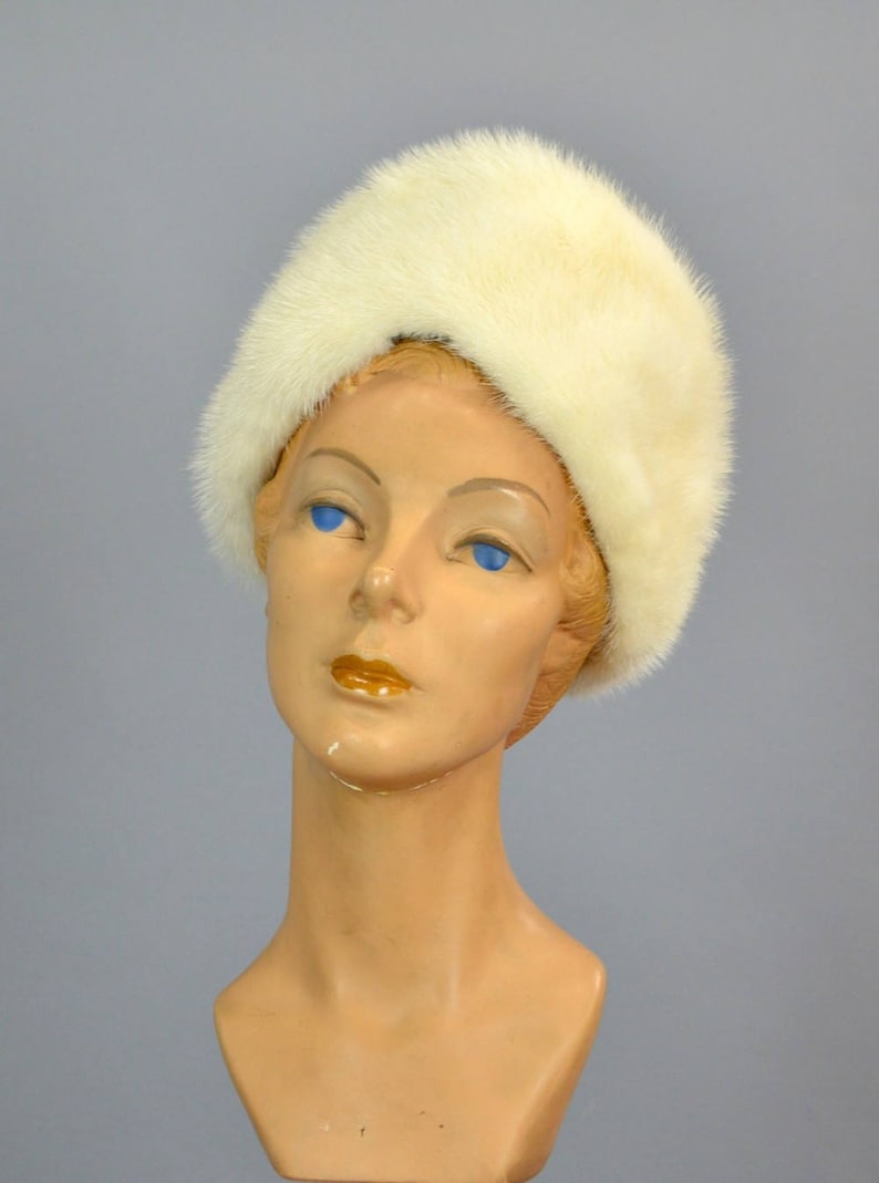 May include: A white fur hat with a wide brim. The hat is worn on a mannequin with a light brown face and blue eyes.