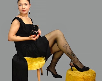 May include: A woman in a black dress and sheer black stockings with a decorative design. She is seated on a yellow velvet stool, wearing black heels, and holding a small black plush toy. The background is a solid gray.