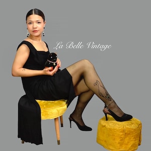 May include: A woman in a black dress and stockings with a decorative design, seated on a yellow stool. She wears black heels and holds a small black plush toy. The image has the text "La Belle Vintage".