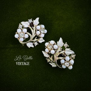 May include: A pair of vintage clip-on earrings. Each earring features a silver-tone metal design with white enamel leaves and clusters of white, pearl-like beads. Clear rhinestones add sparkle. The earrings are set against a dark green background. The text "La Belle VINTAGE" is in the lower left.