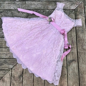 May include: A light pink lace dress with a wide pink satin ribbon tied around the waist. The dress has a scoop neckline and short sleeves. The dress is in a vintage style.