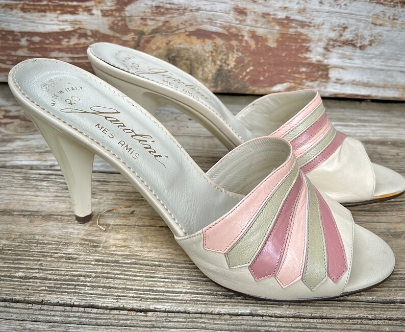 May include: A pair of white leather heeled sandals with pink and green striped detailing. The sandals are labeled "Gianolini Mes Amis" on the insole.