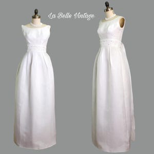 Può includere: Un abito vintage bianco senza maniche con scollatura a barchetta e fascia decorativa in vita. L'abito ha una gonna ampia ed è esposto su un manichino. Il testo "La Belle Vintage" è nell'angolo in alto a sinistra.