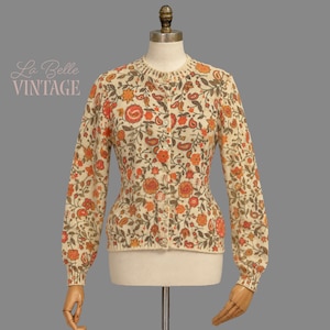 May include: A cream-colored long-sleeve cardigan featuring a repeating floral and paisley pattern in shades of orange, brown, and green. The garment has a round neckline, a front button closure, and ribbed cuffs and hem, displayed on a mannequin.