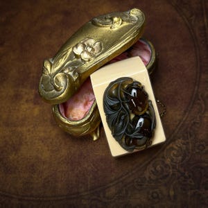 May include: An oval-shaped brooch featuring dark brown, translucent, polished stones set within a carved dark frame, resting on a cream-colored rectangular base next to an ornate, antique-style gold metal trinket box with floral relief.