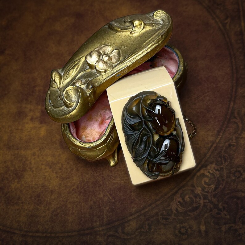 May include: An oval-shaped brooch featuring dark brown, translucent, polished stones set within a carved dark frame, resting on a cream-colored rectangular base next to an ornate, antique-style gold metal trinket box with floral relief.