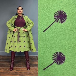 May include: A woman wearing a green coat and skirt with a brown top. The skirt and coat have a pattern of purple dandelions. The woman is wearing brown boots.