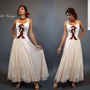 May include: A vintage cream dress with a polka dot pattern and a flowing skirt. The dress has a square neckline, red ribbon accents, and a fitted bodice. The sleeveless dress has a long, elegant silhouette, suitable for a special event. The dress is a classic vintage style.