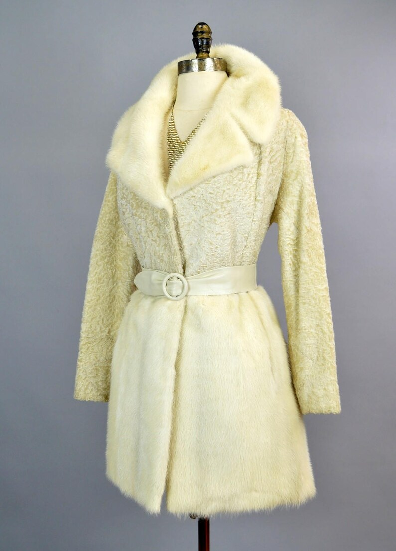 May include: A white fur coat with a large fur collar and a white satin belt. The coat is on a mannequin.