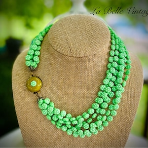 May include: A multi-strand necklace made of green glass beads with a white polka dot pattern. The necklace has a large, round, green and gold button-like clasp.