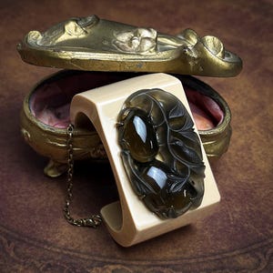 May include: A wide, cream-colored cuff bracelet featuring a large, dark, translucent oval-shaped stone centerpiece with intricate carved leaf and bird details. A thin, metallic chain is attached to the side of the cuff, which rests near a gold-toned, ornate, oval-shaped metal container with a hinged lid.