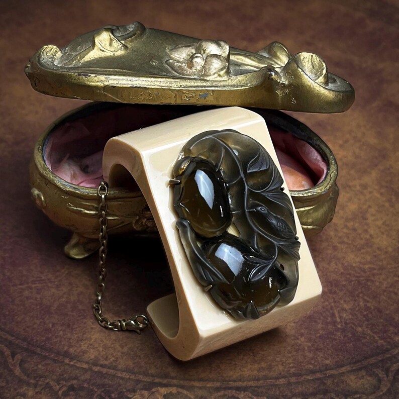May include: A wide, cream-colored cuff bracelet featuring a large, dark, translucent oval-shaped stone centerpiece with intricate carved leaf and bird details. A thin, metallic chain is attached to the side of the cuff, which rests near a gold-toned, ornate, oval-shaped metal container with a hinged lid.
