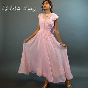 May include: A vintage pink dress with a flowing skirt and a fitted bodice. The dress features a sheer overlay, cap sleeves, and delicate button detailing. The text "La Belle Vintage" is in the upper left corner. The dress is a soft pink color.
