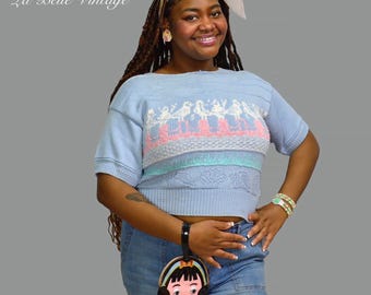 Könnte beinhalten: Hellblauer Vintage-Pullover mit Vogelmuster in Rosa und Weiß. Der Pullover hat kurze Ärmel und einen runden Ausschnitt. Das Bild zeigt auch eine kleine Handtasche mit einem Cartoon-Mädchen-Design und dem Text "La Belle Vintage".