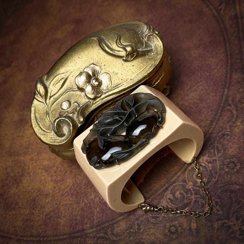 May include: A cream-colored cuff bracelet featuring a dark, translucent carved floral ornament and a delicate metal chain attachment, positioned next to a gold-toned metal object with embossed floral details and a curved, rounded shape.