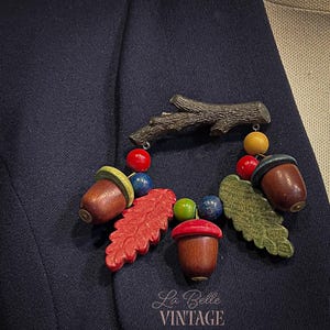 May include: A brooch featuring a dark brown textured branch at the top, from which hang three wooden acorns with brown bodies and colorful caps, two textured oak leaves in red and green, and small round beads in red, blue, yellow, and green.
