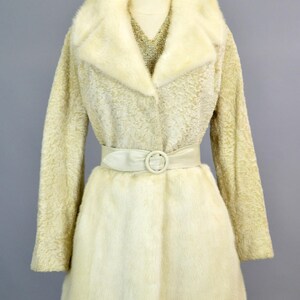 May include: A white fur coat with a large collar and a white leather belt. The coat is on a mannequin.
