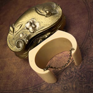 May include: A cream-colored cuff bracelet with a thin metal chain connecting the two ends, positioned next to an ornate, antique-style brass box featuring embossed floral and leaf patterns on its curved, rounded lid and sides.