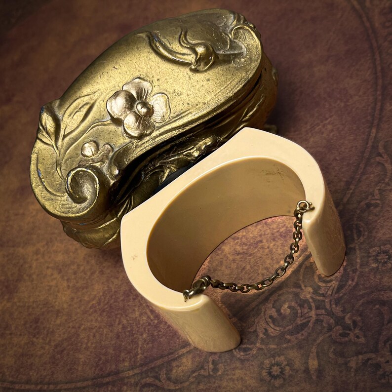 May include: A cream-colored cuff bracelet with a thin metal chain connecting the two ends, positioned next to an ornate, antique-style brass box featuring embossed floral and leaf patterns on its curved, rounded lid and sides.