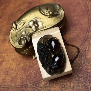 May include: A rectangular cream-colored object features an oval dark brown translucent inlay with carved leaf and bird motifs. A thin metal chain is attached to the side. Behind it sits an ornate, weathered brass-toned metal container with floral relief.