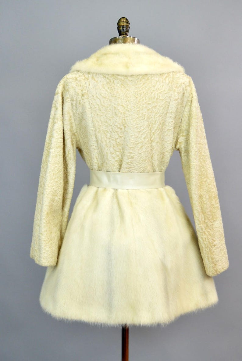 May include: A white faux fur coat with a white satin belt. The coat has a collar and long sleeves.