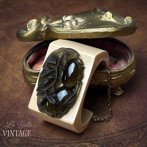 May include: A cream-colored cuff bracelet featuring a dark, translucent, carved floral and bird motif with two smooth, polished, dark brown oval cabochons. The bracelet includes a thin, attached metal chain and rests against a gold-toned metal box.