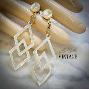 Puede incluir: Un par de pendientes colgantes con recortes geométricos en forma de diamante de nácar blanco iridiscente, suspendidos de pequeños pendientes de perla redonda mediante cadenas de metal dorado. El texto La Belle VINTAGE es visible a la derecha.