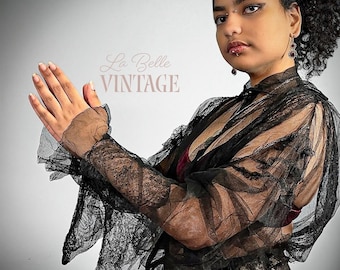 May include: A black sheer lace long-sleeve top featuring a high collar, ruffled cuffs, and intricate floral patterns throughout the fabric. The garment is semi-transparent with a textured finish and includes the text La Belle VINTAGE in the background.