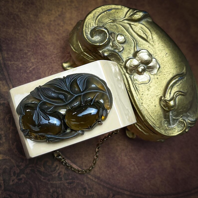 May include: A cream-colored rectangular cuff bracelet featuring a dark brown, translucent, carved amber-like ornament depicting a bird and foliage. Beside it sits a curved, antique-style brass box with embossed floral and leaf motifs.