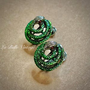 May include: A pair of vintage snake-shaped earrings. The earrings feature a coiled snake design in green with silver-colored snake heads and red accents. The text "La Belle Vintage" is visible in the image.