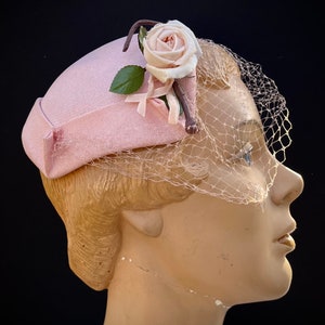 Peut inclure: Un chapeau pillbox rose clair avec un voile délicat. Le chapeau est orné d'une rose rose pâle, d'une feuille verte et d'un petit ruban rose. Le chapeau est présenté sur une tête de mannequin sur fond noir.