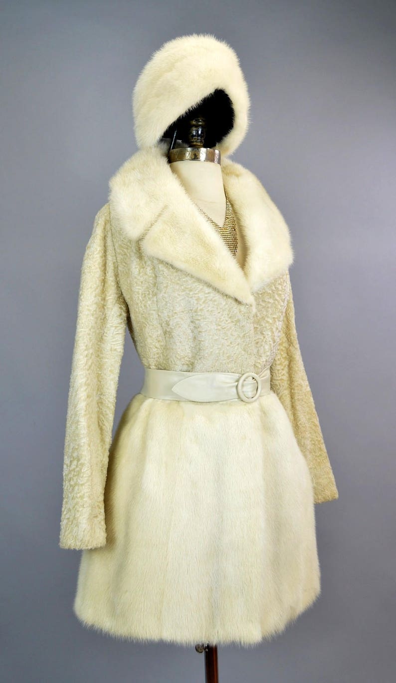 May include: A white fur coat with a matching fur hat and a white leather belt. The coat has a long, flowing style and a large collar.
