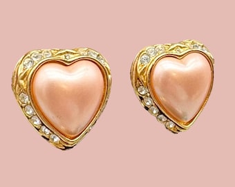 Op de afbeelding: Een paar hartvormige oorbellen. Elke oorbel heeft een lichtroze, parelmoer hart in het midden, omgeven door een goudkleurig frame met heldere kristallen accenten. De oorbellen staan tegen een zachtroze achtergrond.