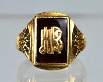 May include: A gold ring featuring a rectangular black onyx inlay centered with a raised gold monogram of the letters HR. The sides of the ring band display intricate, textured relief detailing that extends from the central setting.