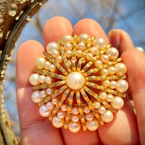 Könnte beinhalten: Goldfarbene Brosche mit Sonnenstrahlendesign, mit einer großen zentralen Perle, umgeben von kleineren Perlen. Die Brosche hat ein detailliertes, verziertes Design, mit goldenen Strahlen, die sich vom Zentrum aus erstrecken. Die Perlen sind cremeweiß.