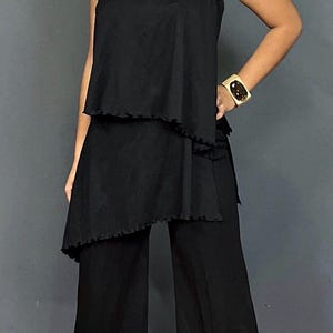 May include: A black one-shoulder top with a ruffled hem and a gold tiger brooch on the shoulder strap, paired with matching black wide-leg trousers. The outfit includes a wide gold cuff bracelet with a dark center stone and gold earrings.