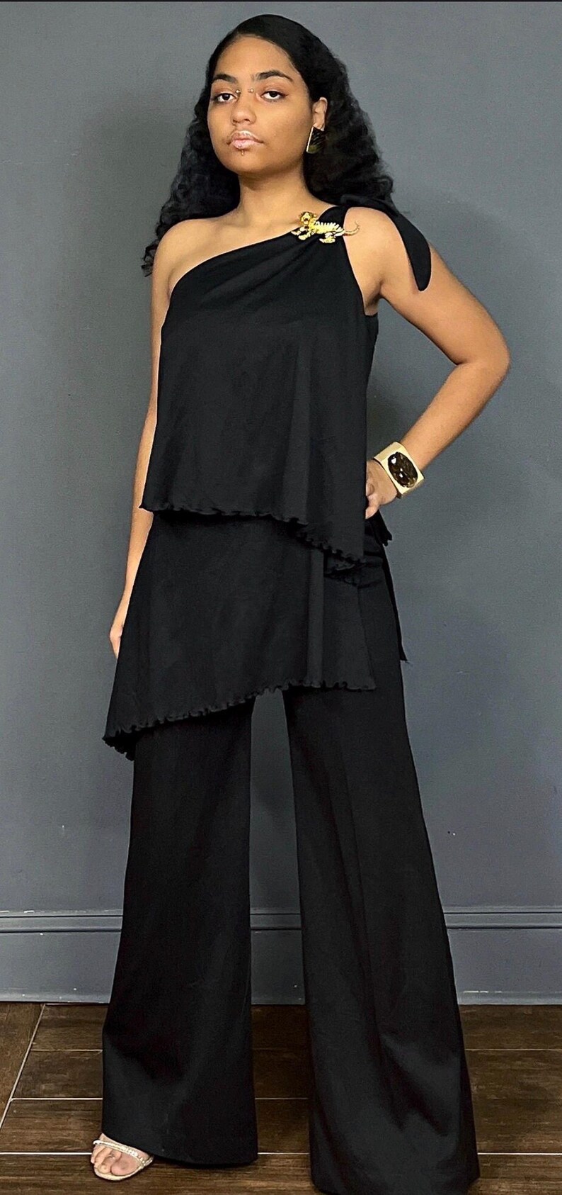 May include: A black one-shoulder top with a ruffled hem and a gold tiger brooch on the shoulder strap, paired with matching black wide-leg trousers. The outfit includes a wide gold cuff bracelet with a dark center stone and gold earrings.