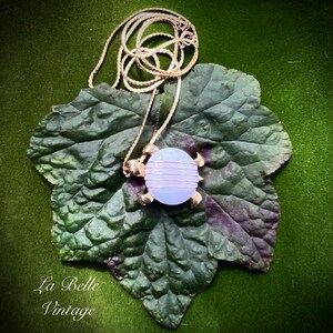 May include: A gold-toned turtle pendant necklace with a light blue stone center wrapped in copper wire. The necklace rests on a large green leaf with a dark green background. The text "La Belle Vintage" is in the lower left corner.