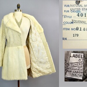 May include: A white fur coat with a shawl collar. The coat is lined with a white fabric. The label on the coat reads "FUR: NATURAL MINK, UNITED STATES FUR ORIGIN: 401, STYLE TOURMALINE, COLOR TOURMALINE, ITEM NO 01402, 179 RN". A smaller label reads "LABEL, CONSUMER, MANUFACT, AIR LABOR, N690".